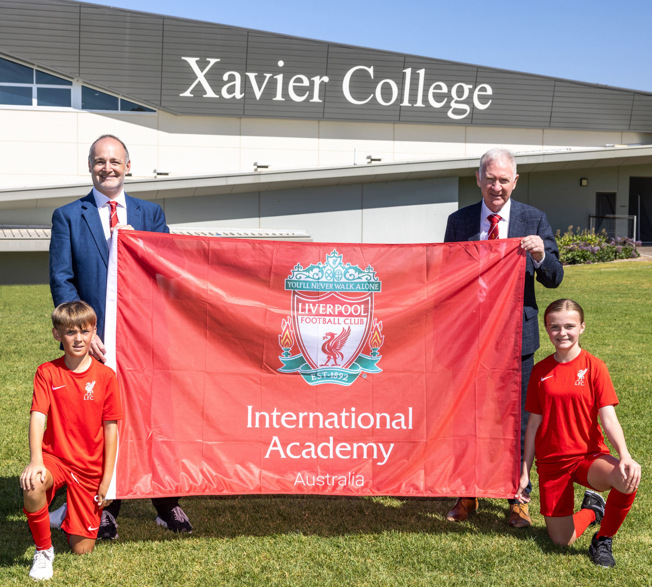 Kevin Kalinko, Director of LFC International Academy and Australian College of Physical Education, and Mark Flaherty, Principal of Xavier College, holding the Liverpool FC International Academy flag, accompanied by two students wearing LFCIA uniforms.