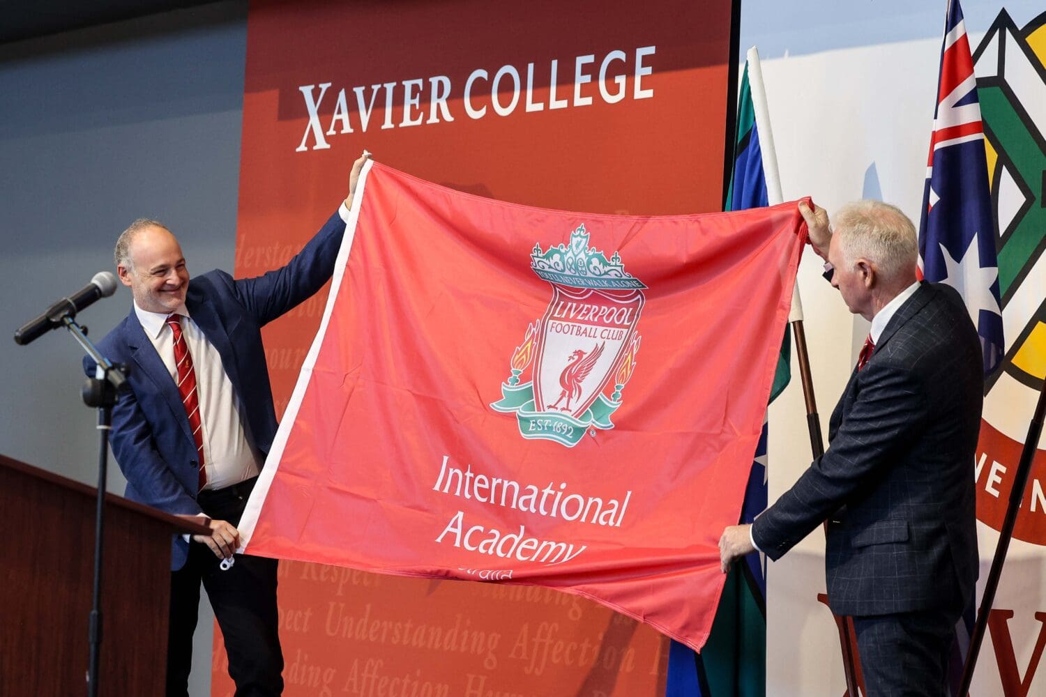 Kevin Kalinko, Director of LFC International Academy, and Mark Flaherty, Principal of Xavier College, standing on stage holding a Liverpool International Academy flag between them.